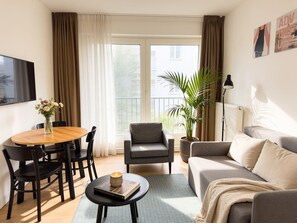 Ein Wohnzimmer mit Sofa, Sessel, Esstisch, Stühlen, einem Couchtisch, einer Zimmerpflanze und einem Fernseher.