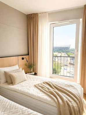 Ein Hotelzimmer mit einem Bett, einem Nachttisch mit Lampe, Blick auf ein Gebäude durch ein Fenster und einem Balkon mit Geländer.