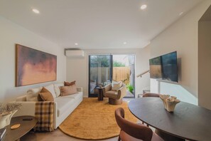 A modern living room with a white sofa, a coffee table, and a dining table with chairs. There is a large painting on the wall and a television mounted on the wall. - Modern 1BR Near Christchurch CBD & Cashel Street (Christchurch)