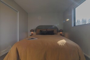 Ett sovrum med en säng, två sänglampor, ett fönster och utsikt över träd.