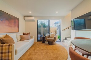 A modern living room with a white sofa, a brown armchair, and a coffee table. There is a large painting on the wall, a flat-screen TV mounted on the wall, and a sliding glass door leading to an outdoor area. - Modern 1BR Near Christchurch CBD & Cashel Street (Christchurch)