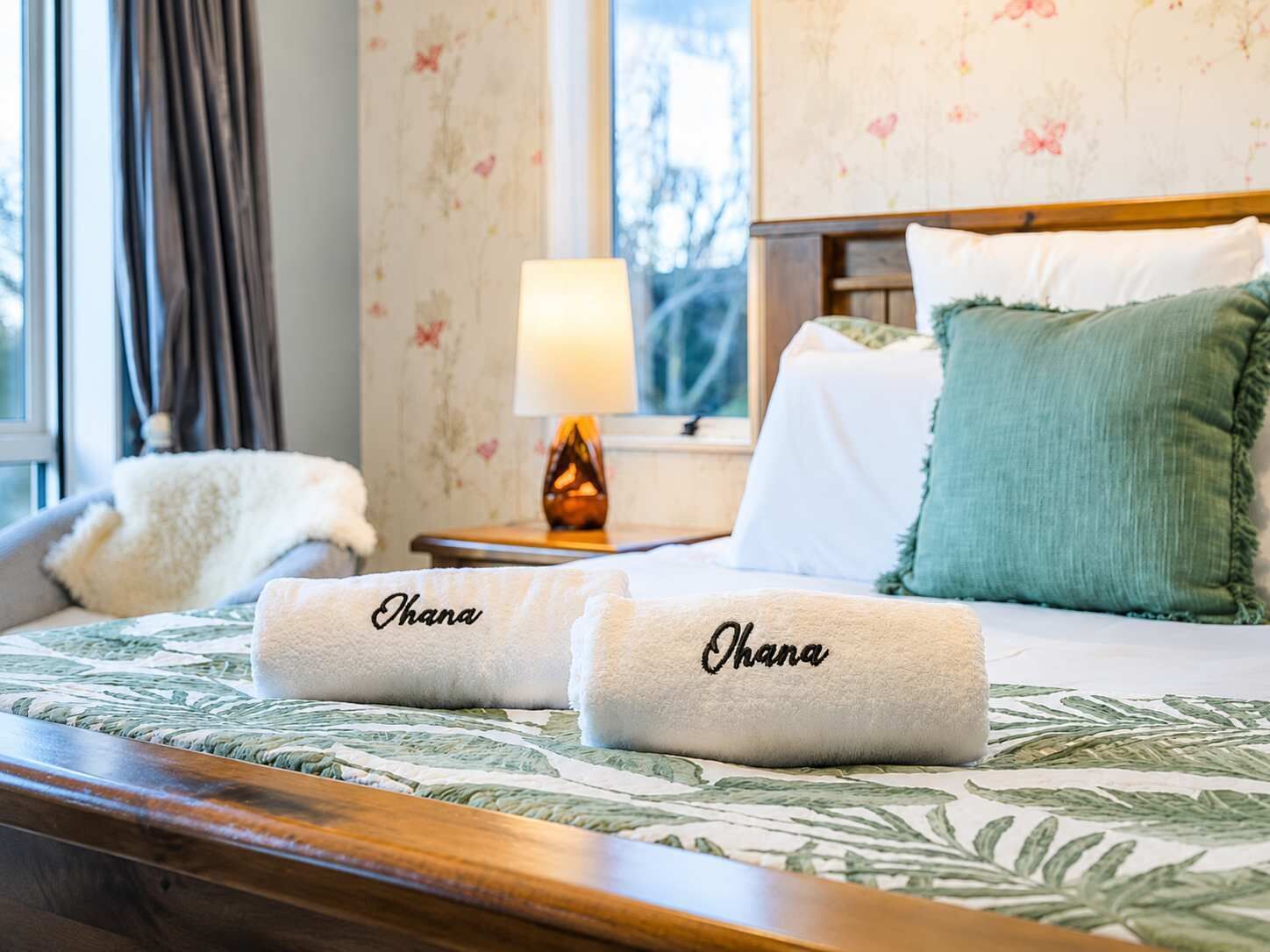 A neatly made bed with two towels labeled 'Ohana' and a nightstand with a lamp.