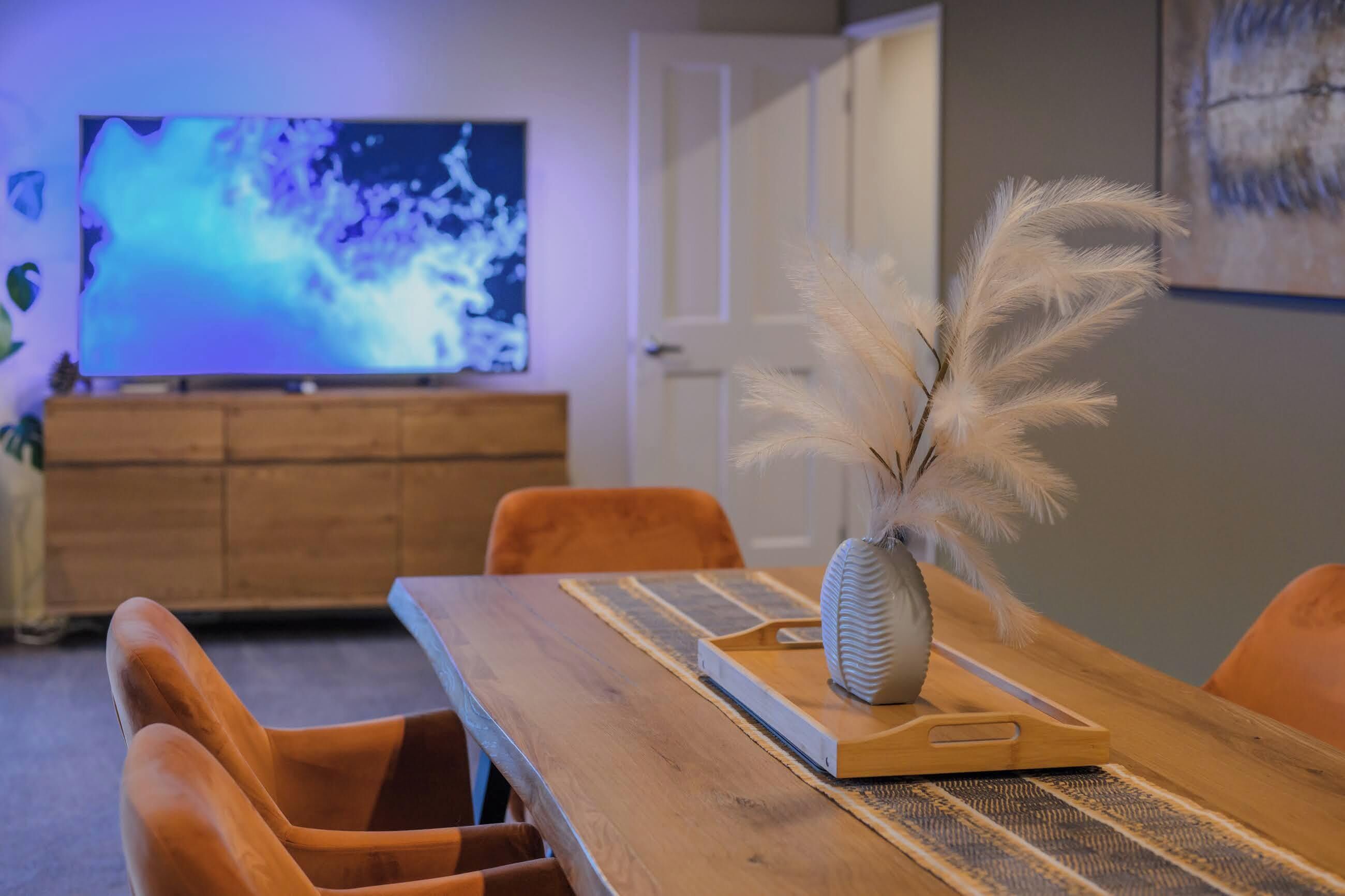A dining area with a wooden table, chairs, a vase with feathers, and a television in the background.