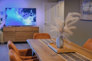A dining area with a wooden table, chairs, a vase with feathers, and a television in the background. - Spacious 4BR Retreat | Halswell Domain Park & Lake (Christchurch)
