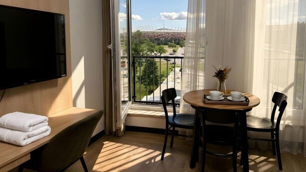 Habitación de hotel con un televisor grande, un escritorio de madera con un mueble para televisión, una mesa redonda con sillas y un balcón con vistas a la ciudad.