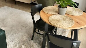 Una sala moderna con un televisor de pantalla plana montado en la pared, una mesa de comedor de madera con individuales tejidos, y una estantería empotrada con plantas.
