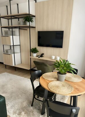 Ein modernes Wohnzimmer mit einem an der Wand befestigten Flachbildfernseher, einem Holzespeisetisch mit gewebten Untersetzern und einer eingebauten Regaleinheit mit Pflanzen.