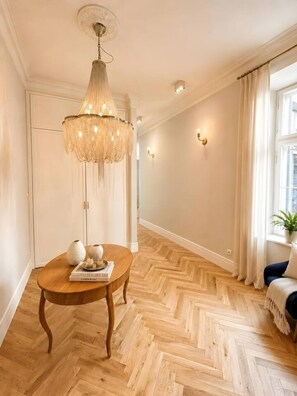 Ein Zimmer mit Holzboden im Fischgrätmuster, einem runden Holztisch mit Büchern und Vasen, einer hängenden Kristallleuchter und einem Fenster mit Vorhängen.
