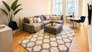 Moderni olohuone, jossa on sohvaryhmä, ruokapöytä tuoleineen, suuri ikkuna verhoilla sekä koristeellinen kattovalaisin.