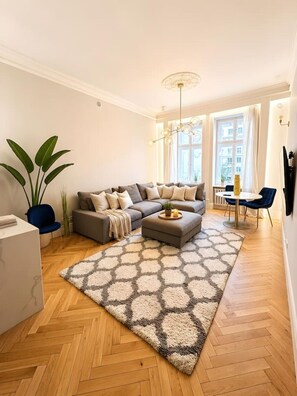 Ein modernes Wohnzimmer mit einer Sofagarnitur, einem Esstisch mit Stühlen, einem großen Fenster mit Vorhängen und einer dekorativen Deckenleuchte.