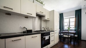 Eine moderne Küche mit Gasherd, Backofen und weißen Schränken. Es gibt eine Essecke mit Tisch und Stühlen, sowie ein Fenster mit Vorhängen.