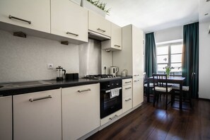 Eine moderne Küche mit Gasherd, Backofen und weißen Schränken. Es gibt eine Essecke mit Tisch und Stühlen, sowie ein Fenster mit Vorhängen.