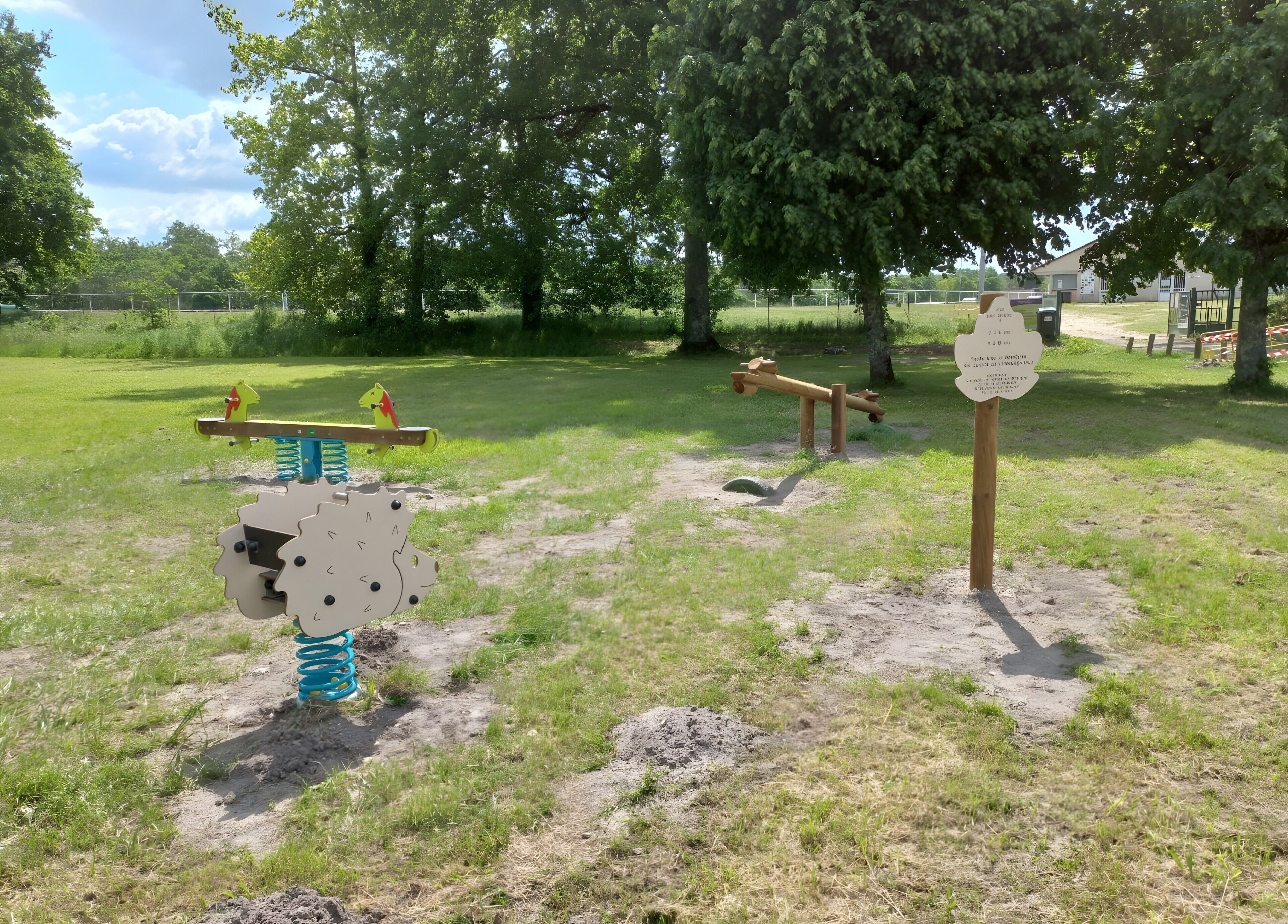 Ein Spielplatz mit verschiedenen Spielgeräten, darunter eine Wippe und ein Karussell, inmitten einer grünen Fläche mit Bäumen im Hintergrund.