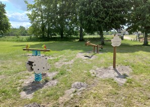 A playground with various play equipment, including a seesaw and a merry-go-round, set in a grassy area with trees in the background.