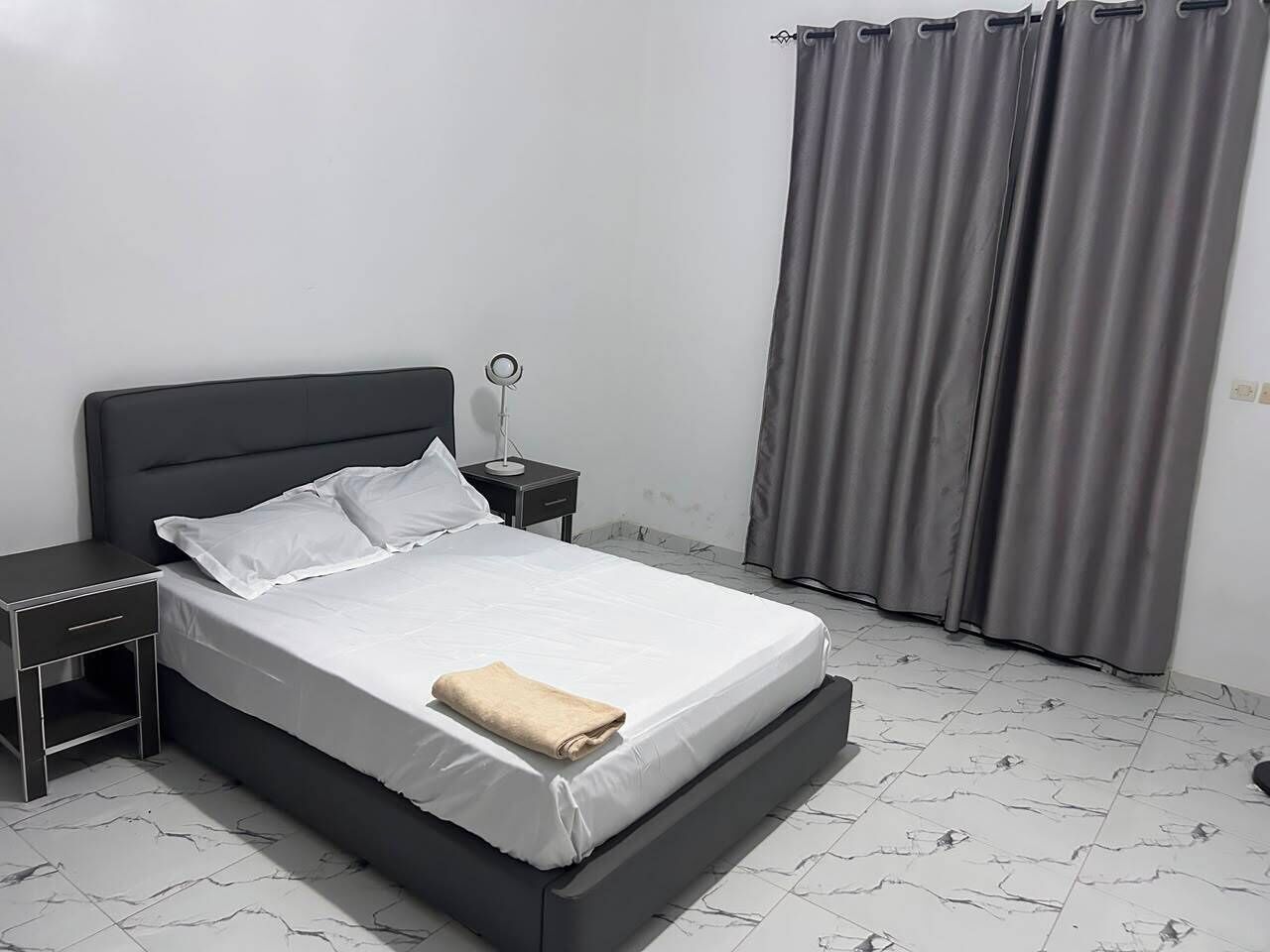 Un lit avec une literie blanche, une table de chevet, une lampe et un rideau gris.