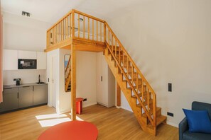A modern living space with a wooden staircase leading to a loft area, a kitchen with a microwave, and a red stool. - Kiez Mezzanine Studio for 4 in Berlin Neukölln (Berlin)
