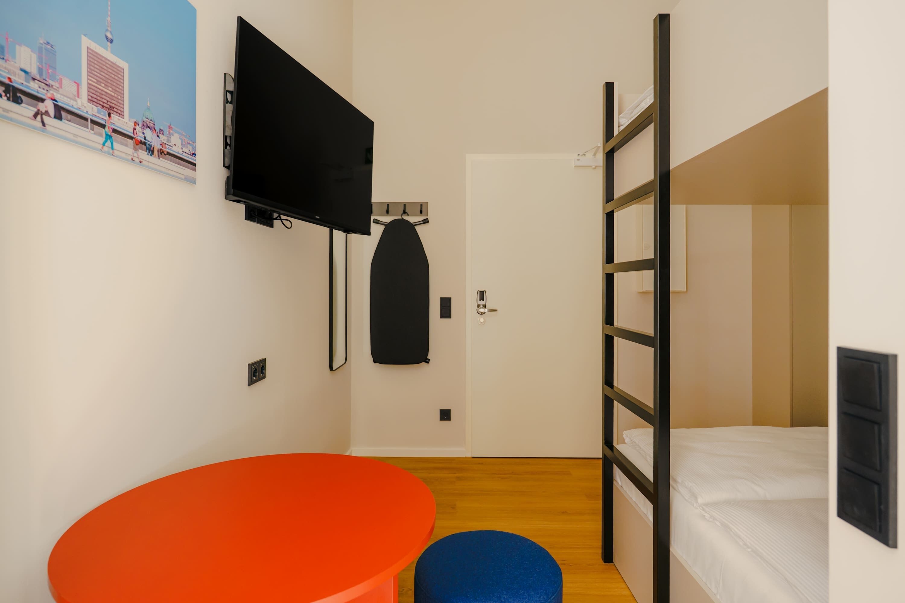 Una habitación compacta con literas, un televisor de pantalla plana en la pared, una pequeña mesa redonda naranja y un reposapiés azul.