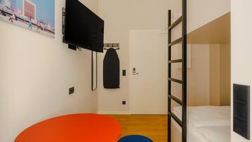Una habitación compacta con literas, un televisor de pantalla plana en la pared, una pequeña mesa redonda naranja y un reposapiés azul.