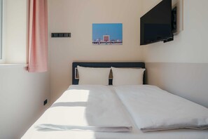 A hotel room with a bed, a television mounted on the wall, and a window with a curtain. - Kiez Studio for 2 Guests in Berlin Neukölln (Berlin)