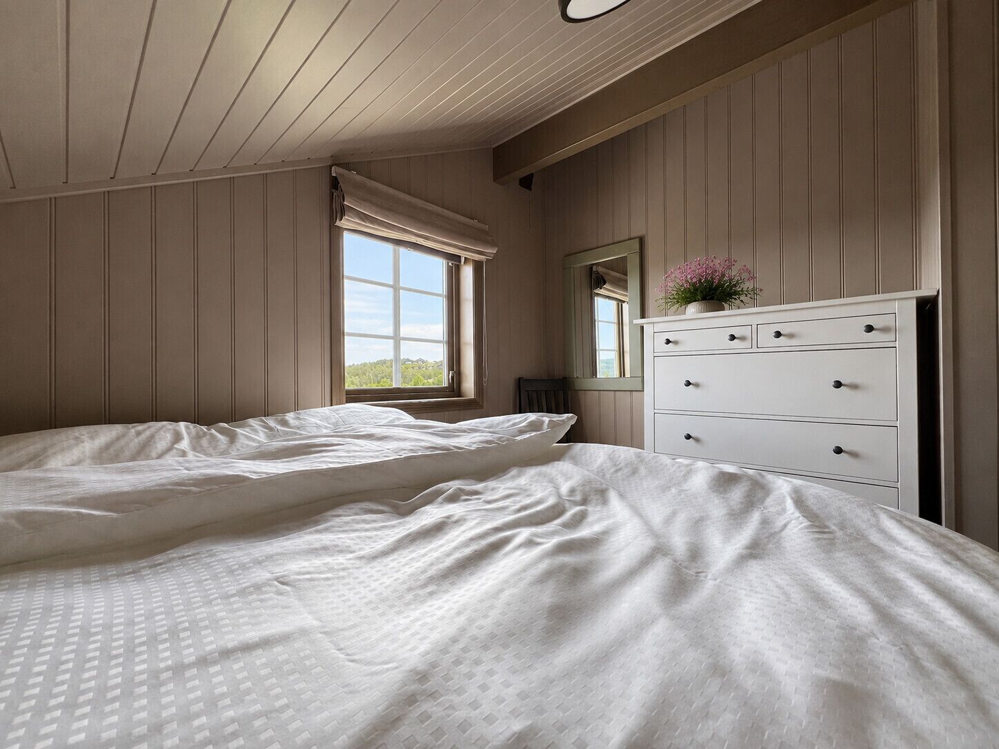 A bedroom with a large bed, a dresser, a window with curtains, and a view of greenery outside.