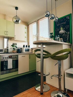 Una cocina con una mesa redonda, taburetes verdes, una pizarra con escritura y una ventana con persianas.