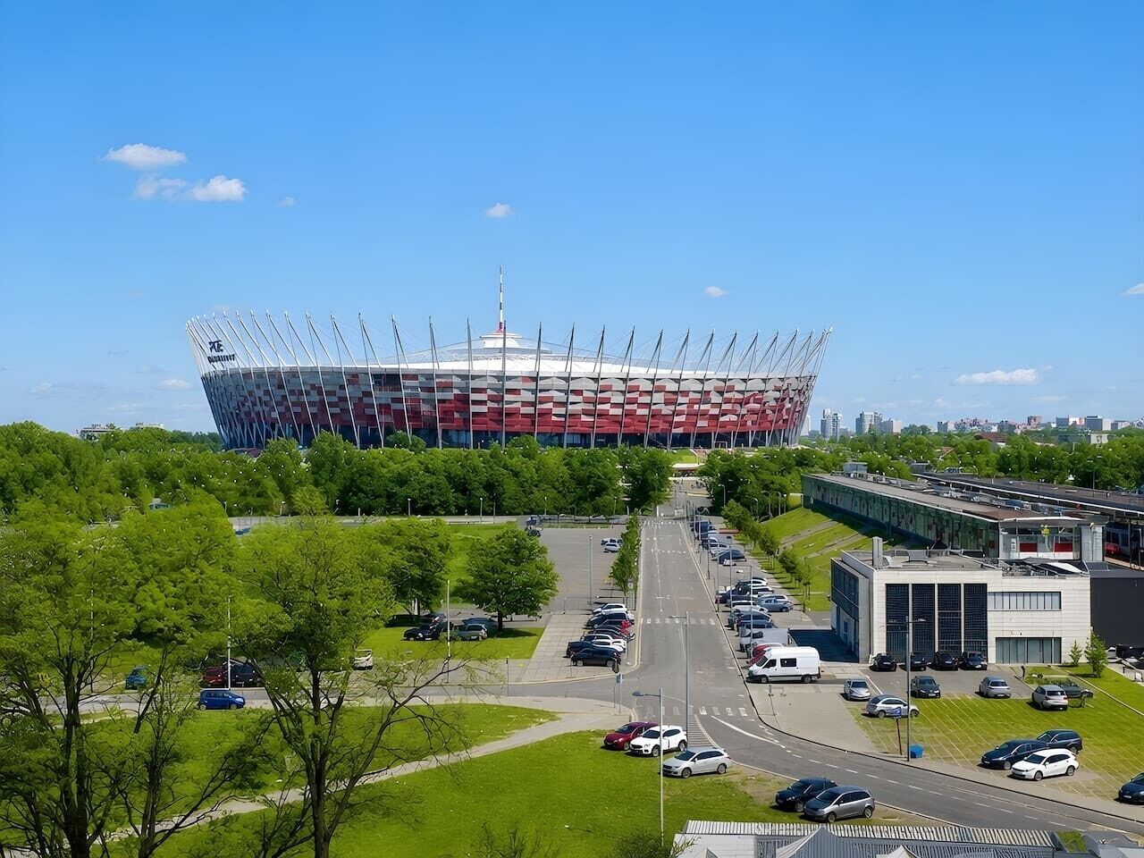 A stadium with a distinctive roof structure, surrounded by parking areas and greenery.