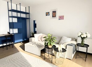 A modern living room with a white sofa, a coffee table, a side table, and a potted plant. There is a blue shelving unit against the wall and a small black table with a vase of flowers. - Old Town Heart - National Stadium Tower 75 (Warsaw)