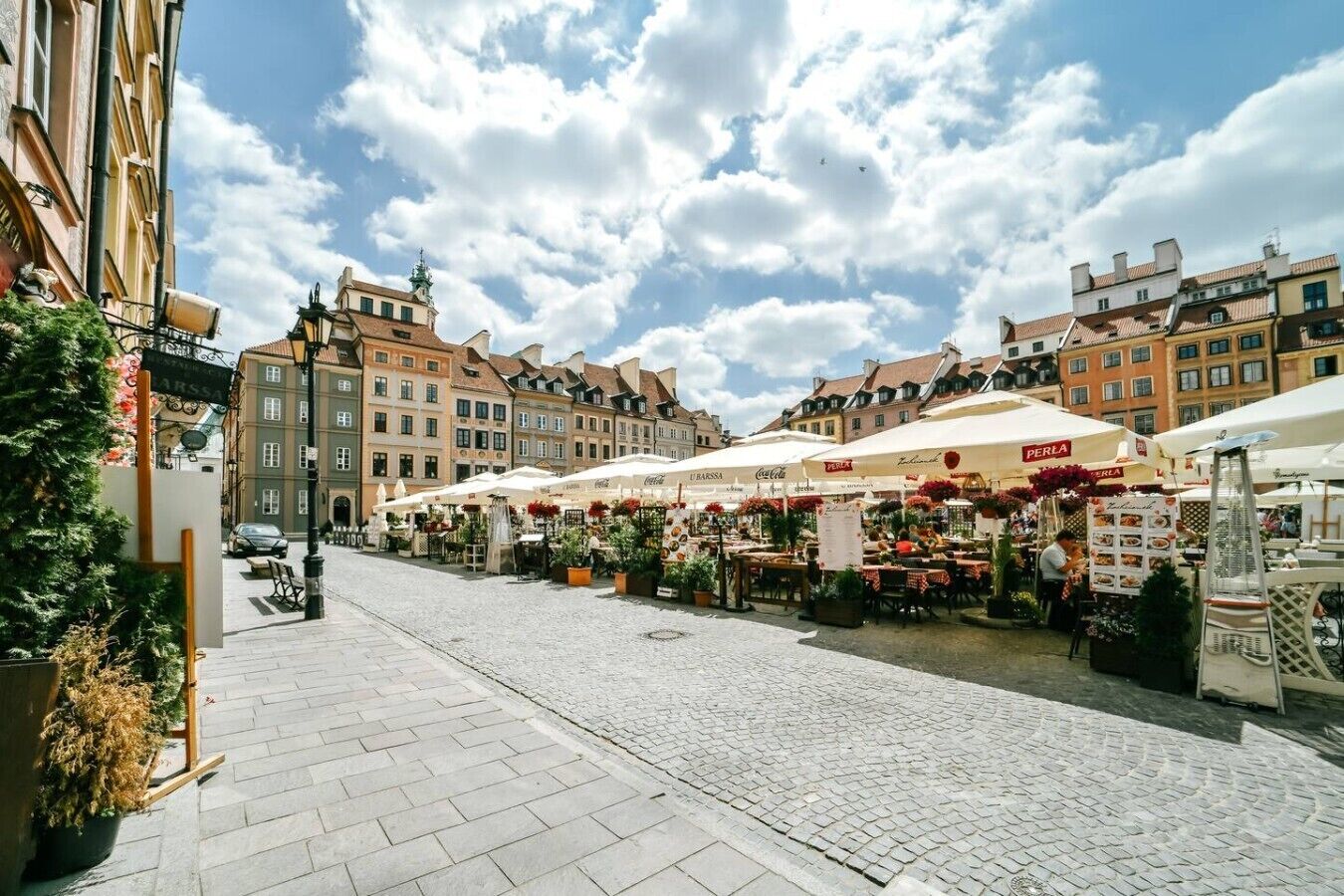 A cobblestone street lined with outdoor cafes and flower stalls, under a clear sky.