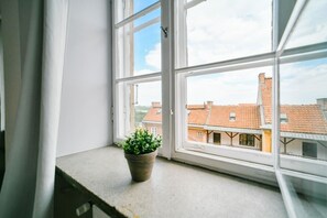 Une fenêtre donnant sur des immeubles, un pot de fleurs posé sur le rebord de la fenêtre et un rideau à moitié tiré.