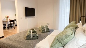 Et hotelværelse med en seng, et fladskærms-tv ophængt på væggen, et spiseområde med stole og et bord, samt et vindue med gardiner.