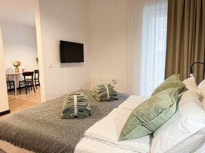 Ein Hotelzimmer mit Bett, einem an der Wand befestigten Flachbildfernseher, einem Essbereich mit Stühlen und einem Tisch sowie einem Fenster mit Vorhängen.