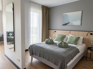 Ein modernes Hotelzimmer mit einem großen Bett, einem Nachttisch mit Lampe, einem Bild an der Wand und Blick ins Freie durch das Fenster.