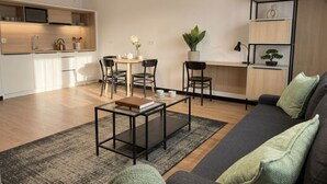 En moderne stue med sofa, sofabord og stole. Der er et køkken med skabe og en mikrobølgeovn. Et bogskab og en potteplante er også synlige.