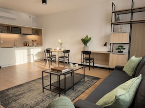 Ein modernes Wohnzimmer mit Sofa, Couchtisch und Sesseln. Es gibt eine Küchenzeile mit Schränken und einer Mikrowelle. Außerdem sind ein Bücherregal und ein Topfpflanze zu sehen.