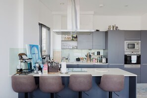 A modern kitchen with a central island, bar stools, a microwave, and an oven. - Stunning 2BDR Flat w Skyline Views in Kentish Town (Greater London)
