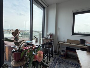 A room with a piano, a table with books and a vase, and a view of the city through large windows. - Stunning 2BDR Flat w Skyline Views in Kentish Town (Greater London)
