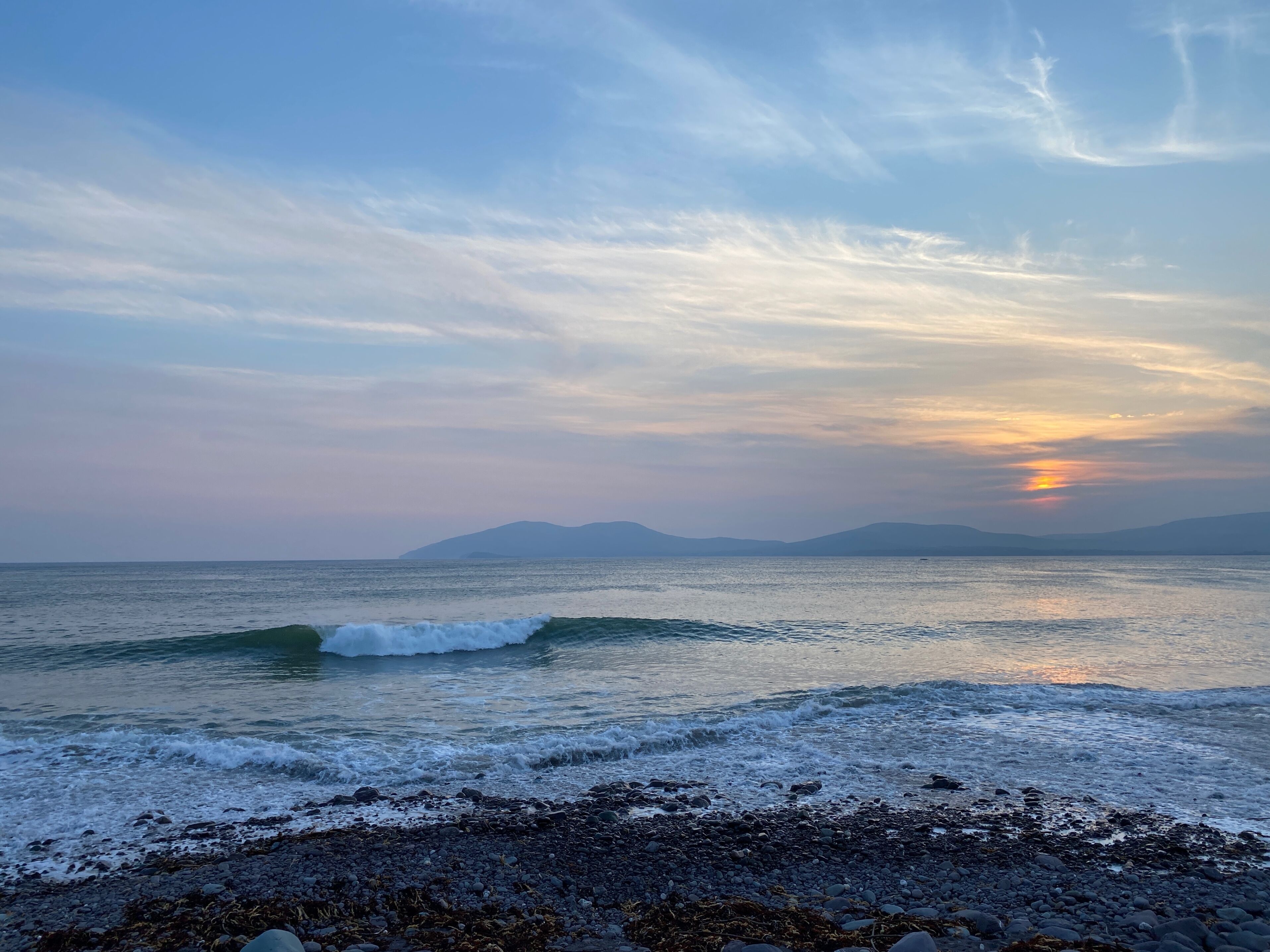 A beach with waves, pebbles, and distant mountains under a clear sky.