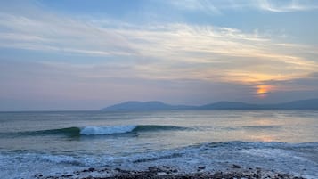 A beach with waves, pebbles, and distant mountains under a clear sky.