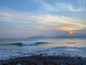 Uma praia com ondas, pedras e montanhas ao longe sob um céu limpo.