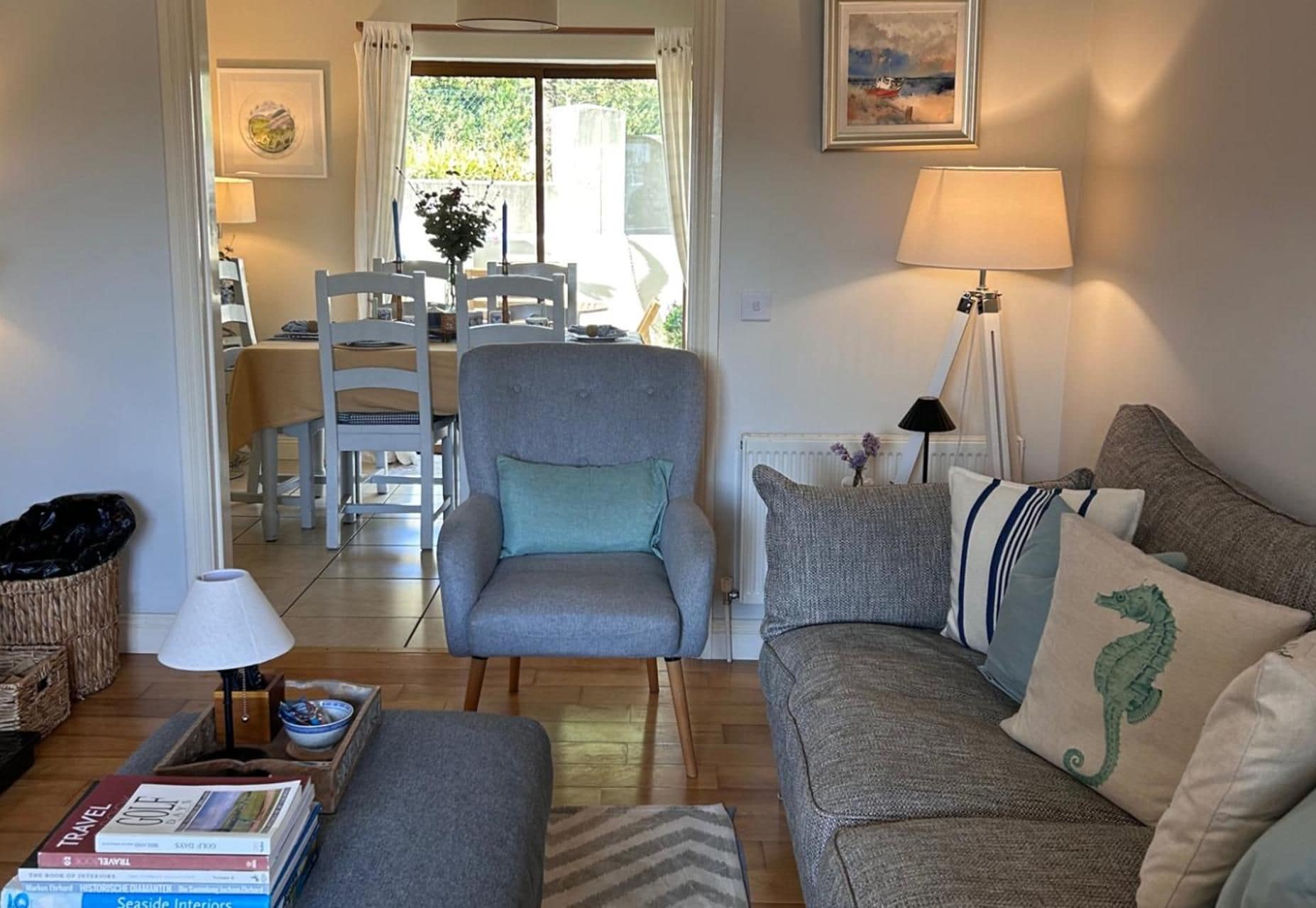 A living room with a grey armchair, a sofa with patterned cushions, a table with books, and a lamp.