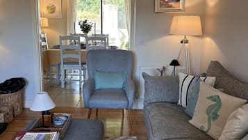 A living room with a grey armchair, a sofa with patterned cushions, a table with books, and a lamp.