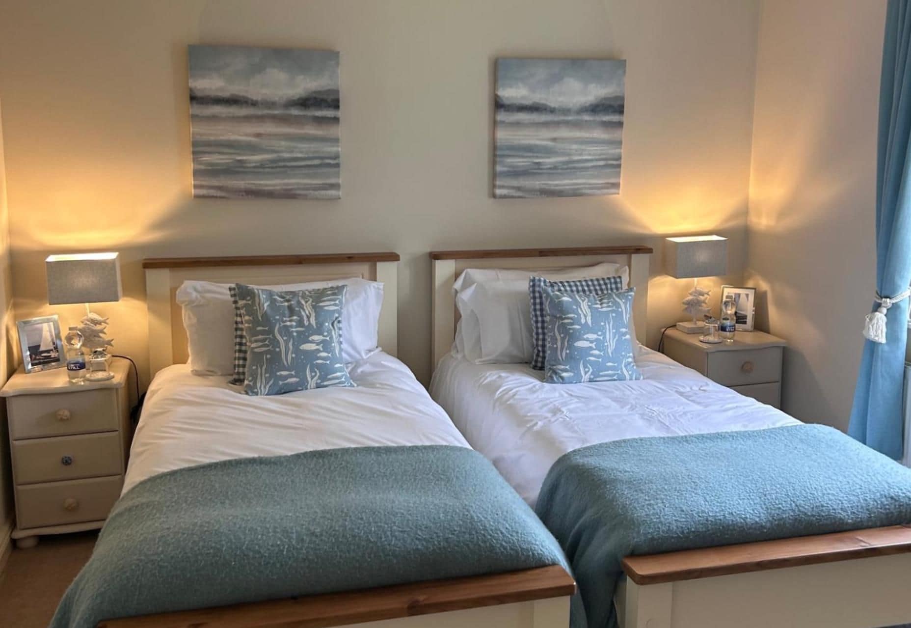 A double room with two beds, each with a nightstand, a lamp, and a small table. There are two wall-mounted paintings above the beds.