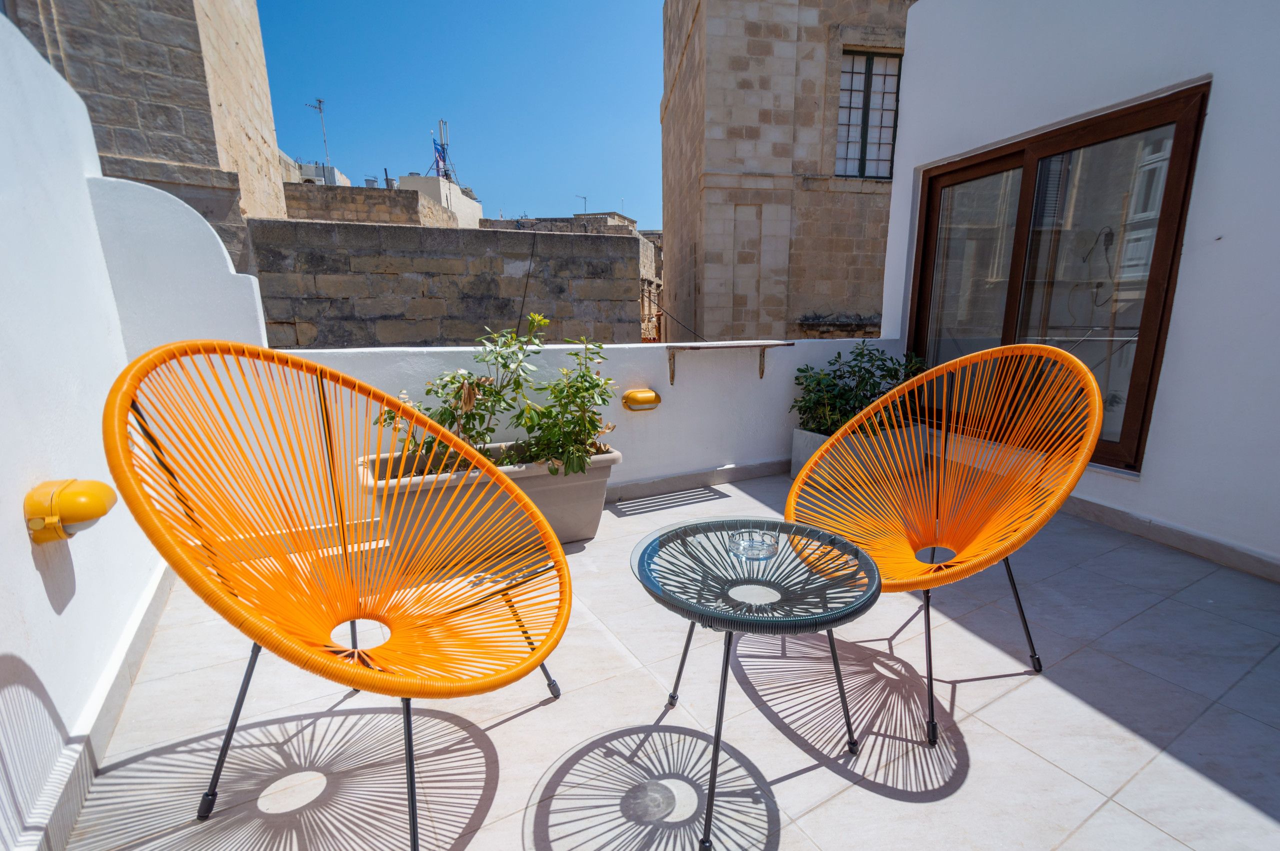 Een terras met twee oranje stoelen en een kleine tafel, een plant in pot en uitzicht op een stadsbeeld.