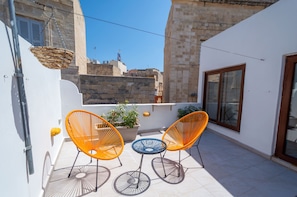 Ein Balkon mit zwei orangefarbenen Stühlen, einem blauen Tisch, einer Topfpflanze und Blick auf ein Steinhaus.