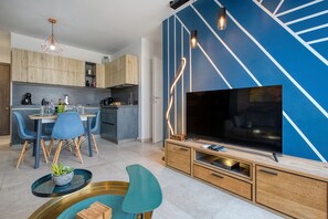 Un salon moderne doté d’un grand téléviseur à écran plat, d’un coin repas avec des chaises bleues et d’une cuisine avec des meubles en bois.