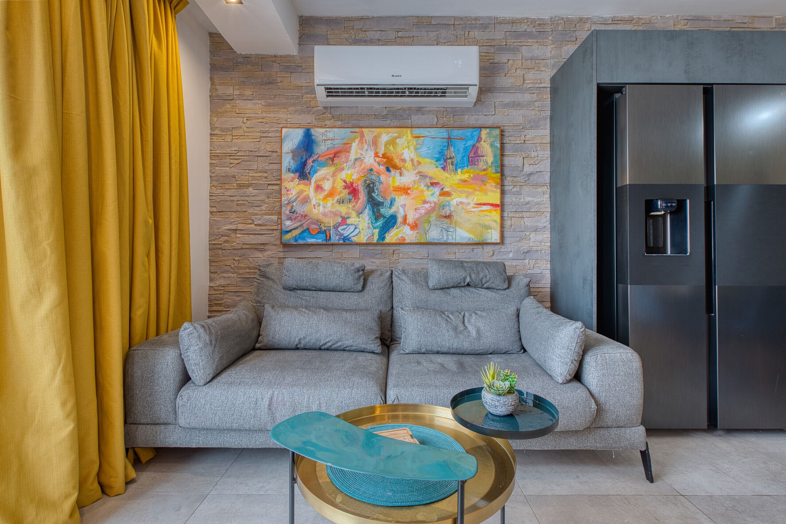 A living room with a grey sofa, a colorful abstract painting, a modern refrigerator, and a small table with a blue cushion.