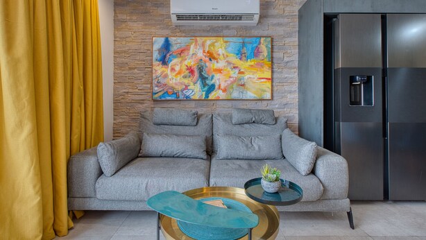 Un salon avec un canapé gris, un tableau abstrait aux couleurs vives, un réfrigérateur moderne et une petite table sur laquelle repose un coussin bleu.