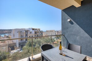 Ein Balkon mit Tisch, einer Flasche und einem Glas, mit Blick auf eine Skyline mit Gebäuden und einem Gewässer.