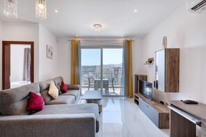 A modern living room with a sofa set, a television, and a balcony with a table and chairs. - Spacious & bright 3 bedroom with balcony KDEB1-1 (Marsaxlokk)