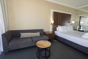 A hotel room with a grey sofa, a wooden stool, a bed with white linens, a nightstand, a lamp, and a large mirror. - Beautiful Apartment on the Gold Coast (Surfers Paradise)